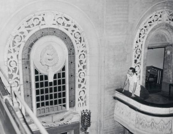 Image Description: Historic photograph of a worker cleaning a balcony a the Studebaker Theater, looking up at one of the decorative drama masks.